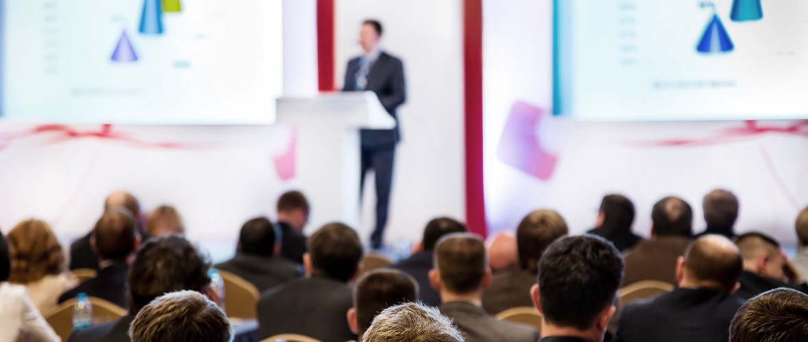 A man stands at the podium in front of a crowd with graphs on a screen behind him