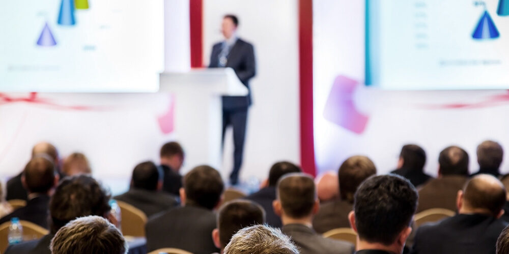 A man stands at the podium in front of a crowd with graphs on a screen behind him