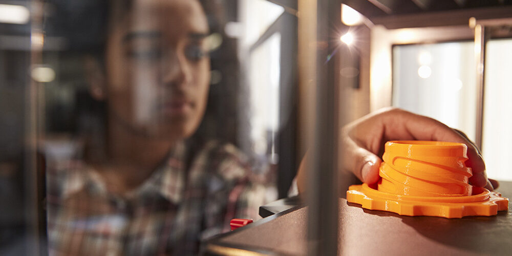 Girl reaching into a machine and holding an orange, gear-shaped part